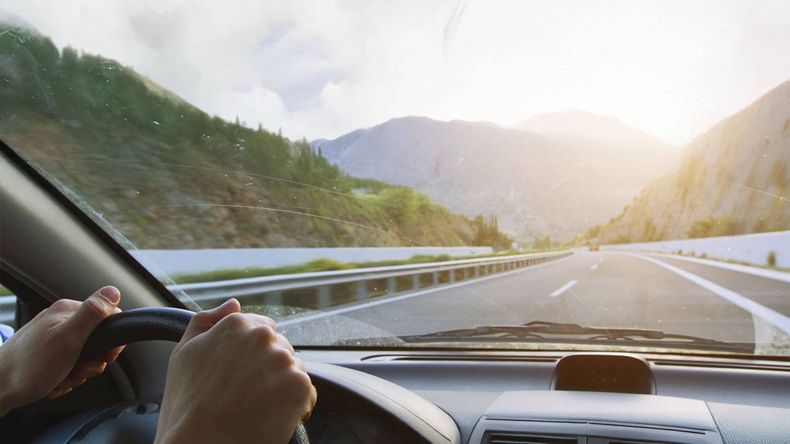 Interior view of a moving car on a mountain highway, hands on the steering wheel, with a landscape of green mountains and a partly cloudy sky